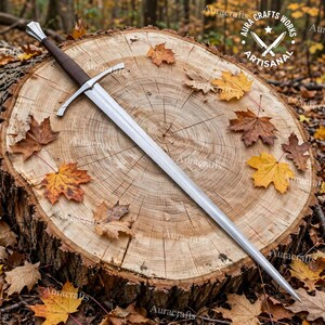 Op de afbeelding: Een stalen langzwaard met een donkerbruine, met leer omwikkelde greep, een zilveren pareerstang en een puntige pommel. Het lemmet is tweesnijdend en loopt uit in een scherpe punt. Een cirkelvormig logo met de tekst Aura Crafts Works Artisanal is zichtbaar.