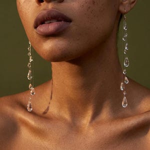 May include: Long drop earrings featuring multiple clear, teardrop-shaped glass beads suspended on thin, transparent filaments. The earrings hang vertically, with the beads arranged in a cascading pattern along the length of the fine, clear thread.