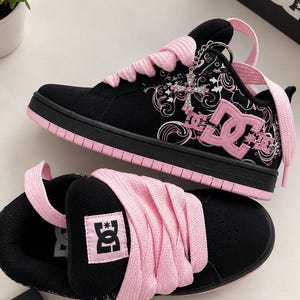 Può includere: Sneaker DC nere e rosa con lacci rosa spessi, un logo DC rosa sul lato e sulla linguetta e un ciondolo a croce argentato attaccato agli occhielli. Il pannello laterale presenta un motivo grafico a vortice bianco e rosa su sfondo nero.