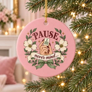 Könnte beinhalten: Ein rundes, rosafarbenes Keramikornament mit einer Hand, die einen Mahjong-Stein hält, umgeben von weißen Hartriegelblüten und grünen Blättern. Der Text PAUSE und never mind... steht auf einem Banner, mit goldener Aufhängung.