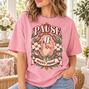 May include: A light pink short-sleeve t-shirt featuring a graphic print of a hand holding a mahjong tile surrounded by flowers and leaves. The text PAUSE is printed above the hand, and the text never mind... is printed on a banner below.