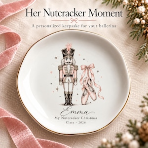 May include: A white ceramic trinket dish with a gold rim featuring a nutcracker illustration, pink ballet slippers, and the text "Her Nutcracker Moment," "Emma," "My Nutcracker Christmas," and "Clara 2026" printed in black and pink.