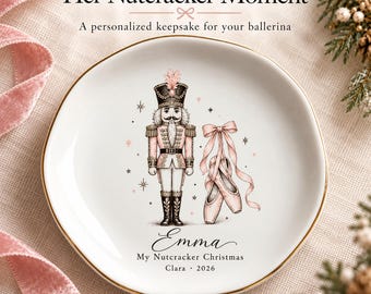 May include: A white ceramic trinket dish with a gold rim featuring a nutcracker illustration, pink ballet slippers, and the text "Her Nutcracker Moment," "Emma," "My Nutcracker Christmas," and "Clara 2026" printed in black and pink.