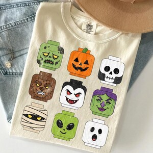 May include: A cream-colored t-shirt featuring a graphic print of nine stylized toy block heads with Halloween-themed designs, including a zombie, pumpkin, skeleton, werewolf, vampire, witch, mummy, alien, and ghost, with a Comfort Colors brand tag.