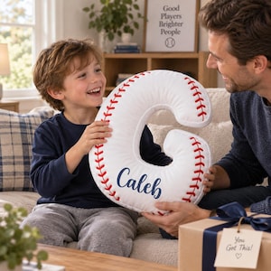 May include: A white plush pillow shaped like the letter C, featuring red stitched baseball-style seams along the edges. The name Caleb is printed in dark blue script on the front of the pillow. The pillow is held by a child and an adult.