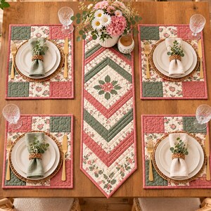 May include: A dining table setting featuring four quilted placemats and a matching table runner in a patchwork pattern of sage green, dusty rose, and cream floral fabric. Each place setting includes a white plate, gold flatware, and a napkin.