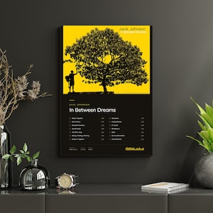 May include: A rectangular wall art print featuring the Jack Johnson album In Between Dreams. The top section displays a black silhouette of a person with a guitar under a large tree against a bright yellow background. The bottom section is black with white text listing the album title and tracklist.
