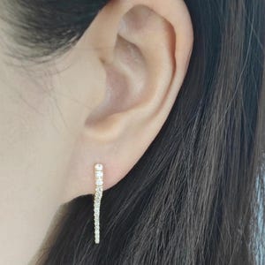 May include: A pair of gold earrings with a single row of small, clear cubic zirconia stones. The earrings are in the shape of a long, thin line.