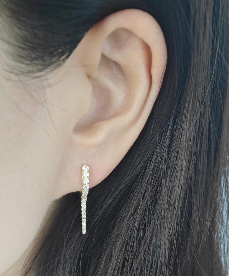 May include: A pair of gold earrings with a single row of small, clear cubic zirconia stones. The earrings are in the shape of a long, thin line.
