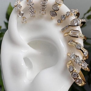 May include: An assortment of gold rings and earrings, each adorned with sparkling diamonds, are displayed on a white ear-shaped form. The jewelry features various diamond cuts and settings, showcasing a range of styles.