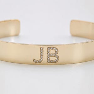 May include: A gold-colored cuff bracelet with the initials "JB" encrusted with small, sparkling stones. The bracelet has a smooth, polished surface and an open design, suitable for various wrist sizes.