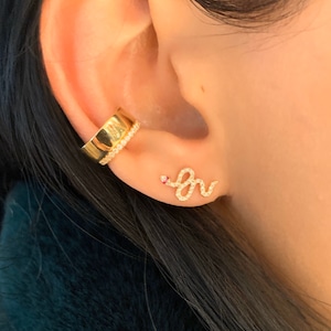 May include: Gold snake earring with a small red stone for the eye. The snake is encrusted with small clear stones. The earring is worn in the upper lobe of an ear.