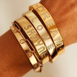 Puede incluir: Una colección de pulseras doradas. Una pulsera presenta un diseño de eslabones de cadena, mientras que otras muestran las palabras "SHAYNE", "LOVE" y "DREAM" en mayúsculas, adornadas con pequeñas piedras brillantes. Las pulseras están apiladas.