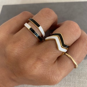 May include: Three gold rings with black and white enamel. The rings are stacked on a finger. The top ring is a simple band with black and white enamel. The middle ring is a chevron shape with black and white enamel. The bottom ring is a simple band with gold metal.