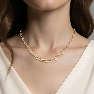 May include: A gold-colored chain necklace with a U-shaped link design. The necklace is worn around the neck and is made of a shiny, reflective metal. The necklace is a statement piece of jewelry.