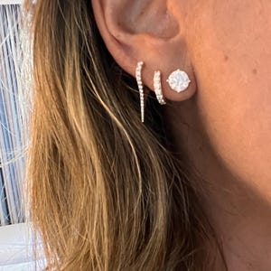 May include: A woman's ear with three diamond earrings. The earrings are a small hoop, a larger round stud, and a long, thin, curved earring.