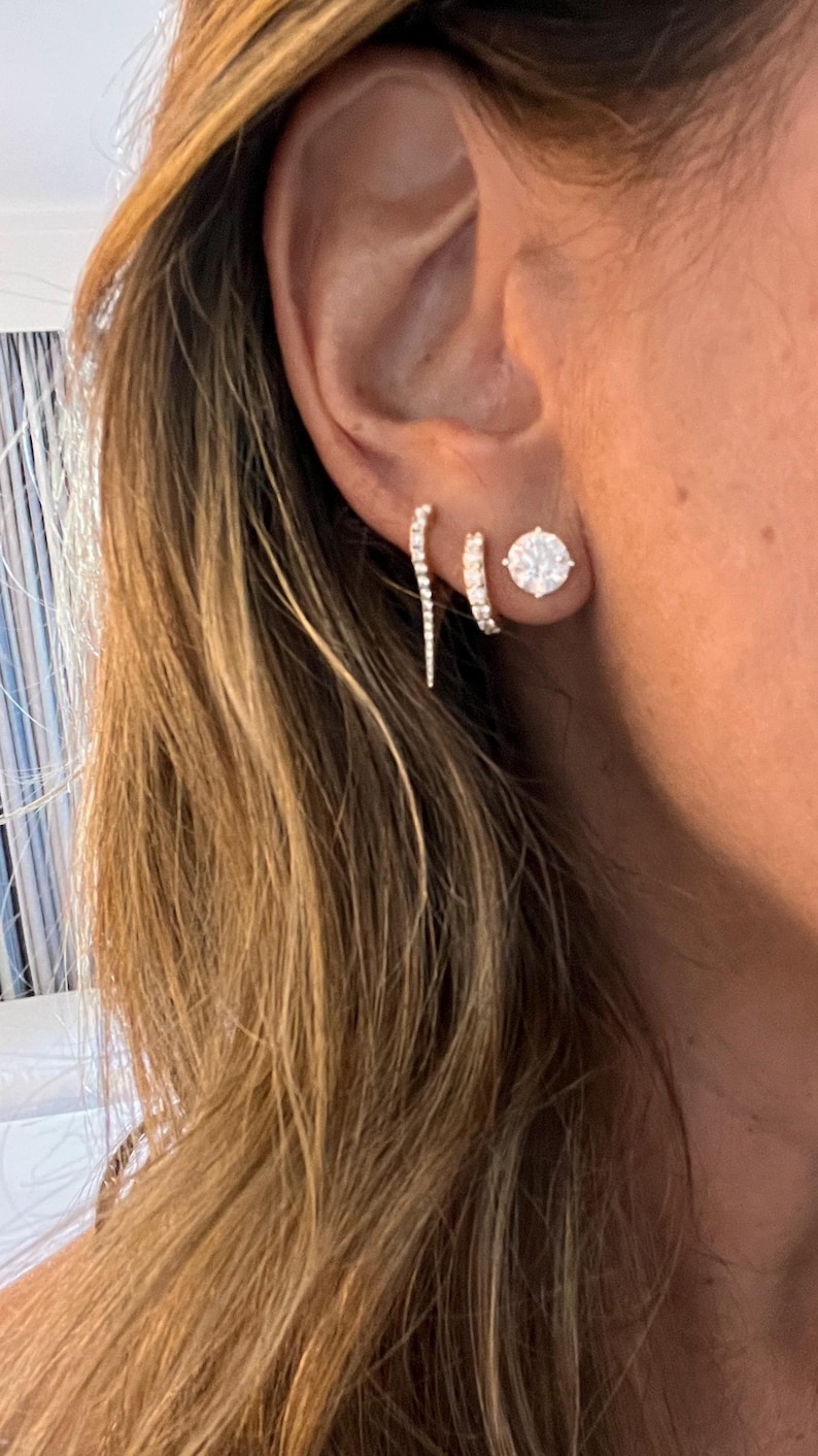 May include: A woman's ear with three diamond earrings. The earrings are a small hoop, a larger round stud, and a long, thin, curved earring.