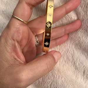 May include: Gold-tone bangle bracelet with star and round crystal accents. The bracelet features a rectangular design with a polished finish. The stars have a black background. A gold ring is also visible.