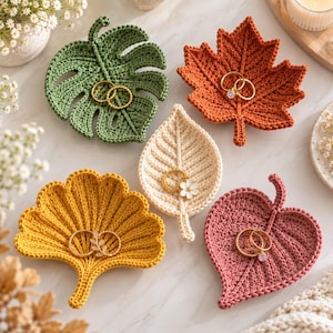 Pode incluir: Cinco pratos de crochê em forma de folha para joias nas cores verde, laranja, creme, amarelo e rosa. Cada prato possui uma nervura central e contém anéis dourados. A coleção inclui uma folha de monstera, uma de bordo e três outras formas.