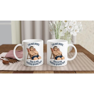 May include: Two white ceramic mugs featuring a cartoon illustration of a person sitting in a striped beach chair with arms crossed. The text "I'm not angry, This is my resting grump face" is printed in dark blue letters above and below the graphic.