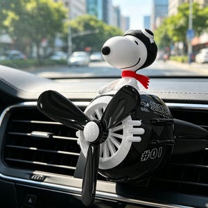 Puede incluir: Un ambientador de coche en blanco y negro con forma de pequeño avión y una figura de Snoopy con gorro de piloto y bufanda roja. La hélice es negra y el cuerpo lleva el texto SNOOPY y #01. Está sujeto a la rejilla de ventilación del vehículo.