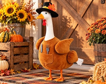 May include: A white goose figurine wearing a black pilgrim hat with a yellow buckle and an orange maple leaf, dressed in a brown plush vest featuring a front pocket that holds a small silver-coloured knife and fork utensil set.