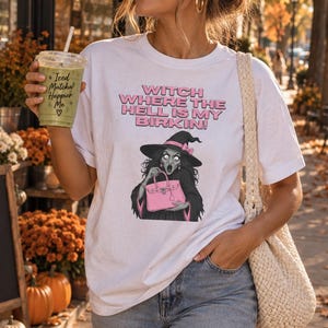 May include: A white short-sleeve t-shirt featuring a graphic of a witch holding a pink handbag. The text on the shirt reads, "WITCH WHERE THE HELL IS MY BIRKIN!". The design is printed in pink and black ink on the front of the cotton crewneck shirt.