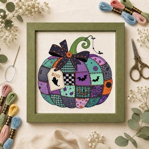 Könnte beinhalten: Ein gerahmtes Kreuzstich-Stickbild mit einem Patchwork-Kürbis in Lila-, Türkis- und Orangetönen. Der Kürbis ist mit einer dunklen Schleife, einem Etikett mit der Aufschrift BOO und einem Geister-Symbol sowie kleinen Fledermaus-Silhouetten verziert.