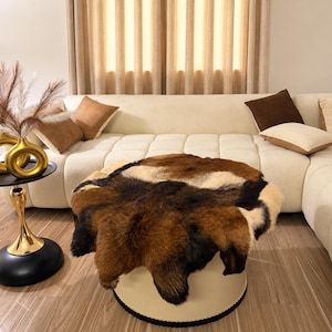 May include: A round ottoman featuring a plush, multi-colored animal hide cover with patches of dark brown, light brown, and cream fur. The base of the ottoman is a solid, smooth cream-colored cylinder with a dark trim along the bottom edge.