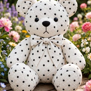 Könnte beinhalten: Ein gesteppter weißer Teddybär mit einem schwarzen Punktemuster auf dem gesamten Körper. Der Bär hat schwarze Knopfaugen, eine schwarz gestickte Nase und einen Mund und trägt eine passende weiße Fliege mit schwarzen Punkten.