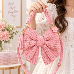 May include: A light pink crochet handbag shaped like a large bow. The bag features a textured knit pattern, a matching top handle, and a detachable shoulder strap with a gold-tone buckle. A pink tassel and a small flower charm are attached to the side.