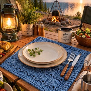 Op de afbeelding: Een vierkante blauwe gehaakte placemat met een herhalend waaiervormig steekpatroon. De placemat ligt op een houten tafel en bevat een beige keramisch bord, een zilveren vork met een houten handvat en een zilveren mes met een houten handvat.