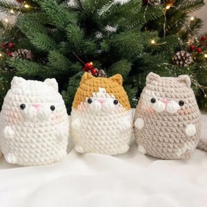 May include: Three round, crocheted cat plushies featuring soft yarn textures. The left cat is solid white, the center cat is white with a tan head, and the right cat is a light brown tabby pattern. Each has black bead eyes and a pink stitched nose.
