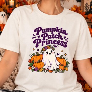 Könnte beinhalten: Ein cremefarbenes Kurzarm-T-Shirt mit einer Grafik eines weißen Geistes, der einen Blumenkranz trägt, umgeben von orangefarbenen Kürbissen und Herbstblättern, mit dem violetten Schriftzug Pumpkin Patch Princess über der Illustration.
