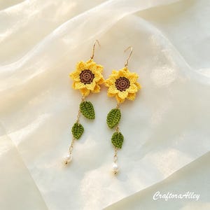 以下が含まれることがあります： 茶色の中心を持つかぎ針編みの黄色いヒマワリの花、緑のかぎ針編みの葉、金色の金属製フックから吊り下げられた小さな白い真珠を特徴とする一対のぶら下がりイヤリング。右下隅にCraftoraAlleyというテキストが表示されています。
