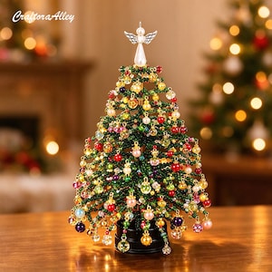 Op de afbeelding: Een decoratieve kerstboom voor op tafel, gemaakt van groene kralen en versierd met diverse kleurrijke glazen en parelachtige ornamenten. Een zilverkleurige engelfiguur staat op de top. De voet is zwart en de tekst CraftoraAlley is zichtbaar.
