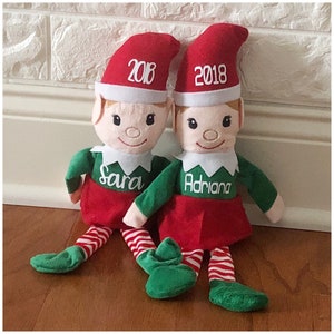 Può includere: Due bambole elfo personalizzate con gambe a strisce rosse e bianche, costumi da elfo verdi e cappelli rossi. I cappelli hanno l'anno 2018 ricamato. Le bambole sono sedute su un pavimento di legno. L'elfo a sinistra ha il nome "Sara" ricamato sul suo costume, e l'elfo a destra ha il nome "Adriana" ricamato sul suo costume.