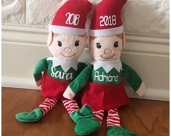 May include: Two personalized elf dolls with red and white striped legs, green elf suits, and red hats. The hats have the year 2018 embroidered on them. The dolls are sitting on a wooden floor. The elf on the left has the name "Sara" embroidered on its suit, and the elf on the right has the name "Adriana" embroidered on its suit.