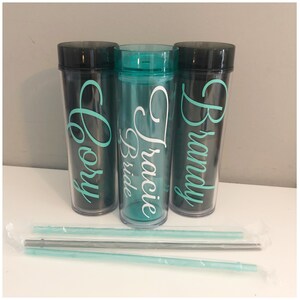 May include: Three clear plastic tumblers with black lids and personalized names. The middle tumbler is teal and says "Tracie Bride". The tumblers are accompanied by three clear plastic straws.
