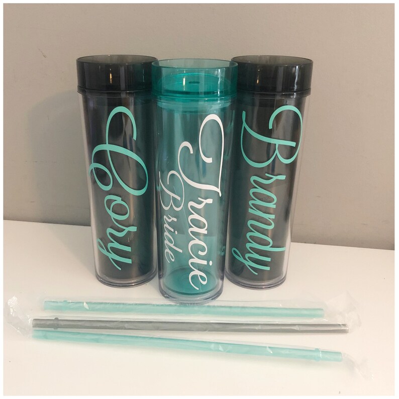May include: Three clear plastic tumblers with black lids and personalized names. The middle tumbler is teal and says "Tracie Bride". The tumblers are accompanied by three clear plastic straws.
