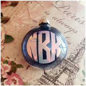 May include: A round, dark blue Christmas bauble with glitter and a silver cap. The bauble features the white letters "MRK" in a monogram design. A silver string is attached for hanging.