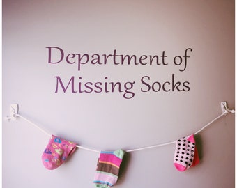 May include: A white string with three socks hanging on it. The socks are pink with white polka dots, pink with white and green stripes, and black with white polka dots. The wall behind the socks has the text "Department of Missing Socks" written on it.