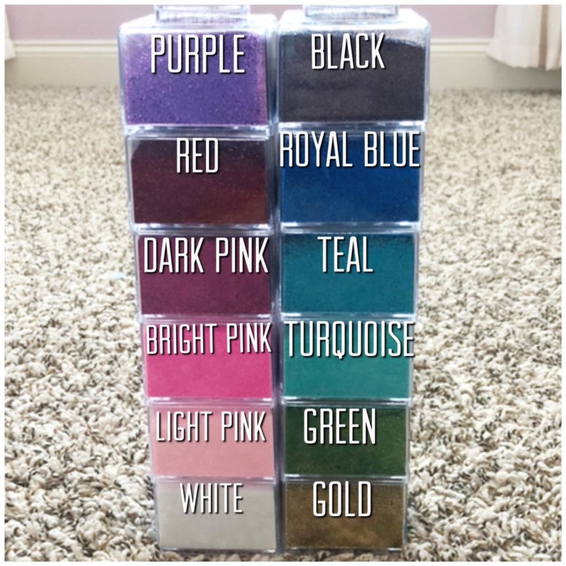May include: A display of clear plastic containers filled with coloured sand. The colours are purple, red, dark pink, bright pink, light pink, white, black, royal blue, teal, turquoise, green, and gold. Each container is labelled with the colour name.