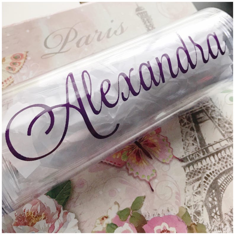 K&ouml;nnte beinhalten: Ein durchsichtiger Plastikbecher mit einem violetten Vinyl-Aufkleber, auf dem "Alexandra" steht.