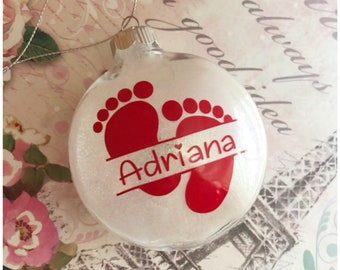 May include: A white glitter Christmas ornament with a red footprint design and the name "Adriana" written on a silver banner.