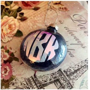 May include: A dark blue Christmas bauble with a silver cap and hanging string. The bauble features a holographic monogram in white letters. The background has a floral design.