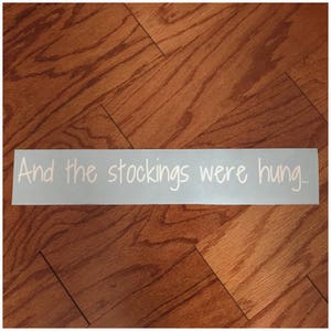 Op de afbeelding: Een witte vinyl sticker met de tekst "And the stockings were hung..." op een lichtblauwe achtergrond.