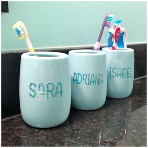 Peut inclure: Trois porte-brosses à dents en céramique bleu clair avec les noms "Sara", "Adriana" et "Isabel" écrits en paillettes turquoise sur le devant.