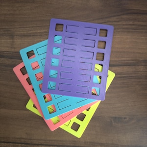 Puede incluir: Una pila de cuatro plantillas rectangulares de plástico en colores morado, verde azulado, coral y amarillo. Cada plantilla presenta una cuadrícula de recortes rectangulares uniformes dispuestos en columnas y filas para marcar.