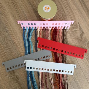 Pode incluir: Um organizador de fios de bordar de plástico rosa carregado com vários tons de fios azuis, castanhos e vermelhos, acompanhado por três organizadores vazios adicionais em vermelho, cinzento e branco, e uma vela redonda com a etiqueta COSY FALL COLLECTION.